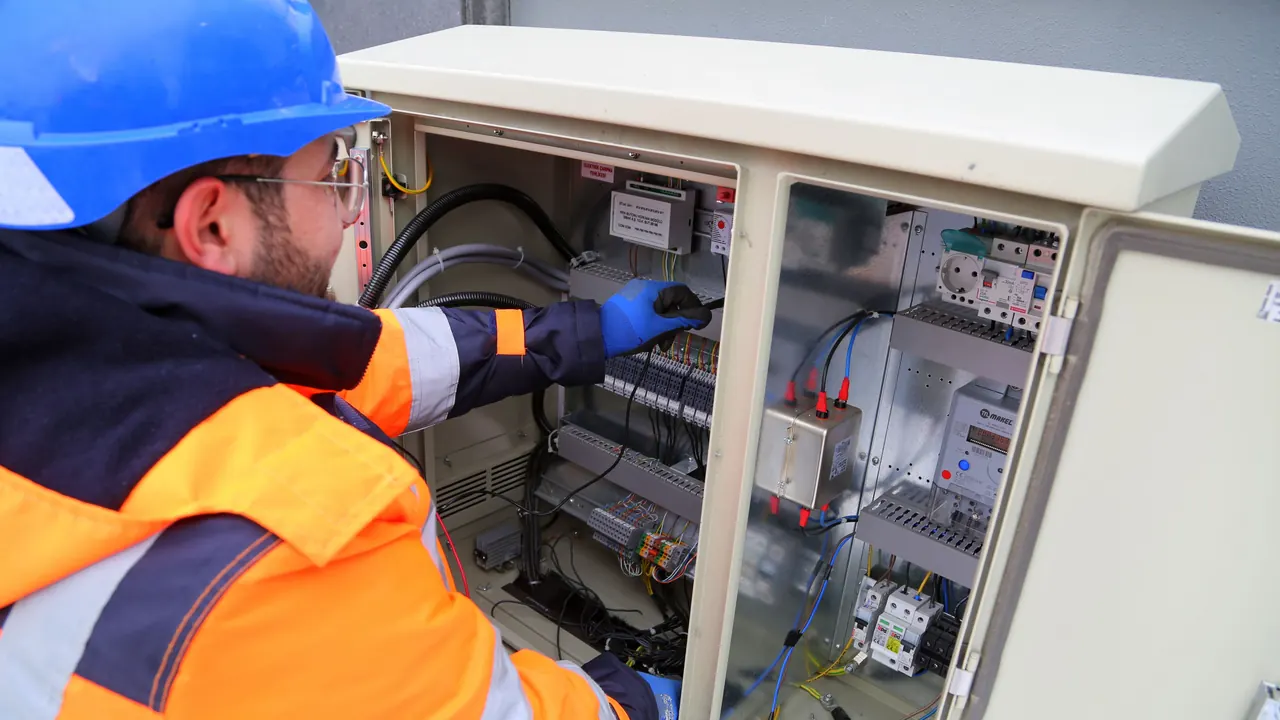 Électricien casqué en intervention sur un tableau électrique, gants et outils visibles.