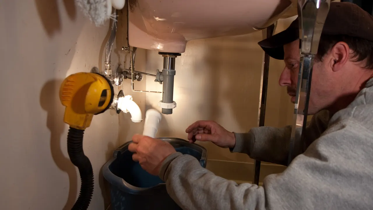 Plombier en dépannage sous un lavabo, siphon démonté et seau pour récupérer l’eau.