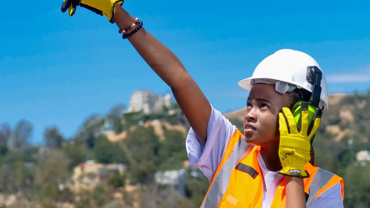 Artisane casquée en gilet orange, au téléphone, indiquant une direction sur un chantier.